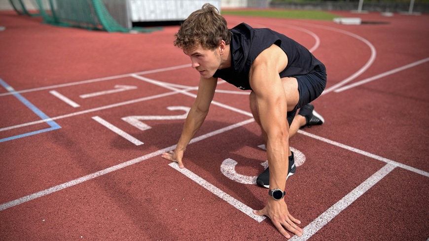 Muž s inteligentnými hodinkami Garmin fēnix® 9 Pro na zápästí sa v bloku na atletickom ovále pripravuje na šprint.