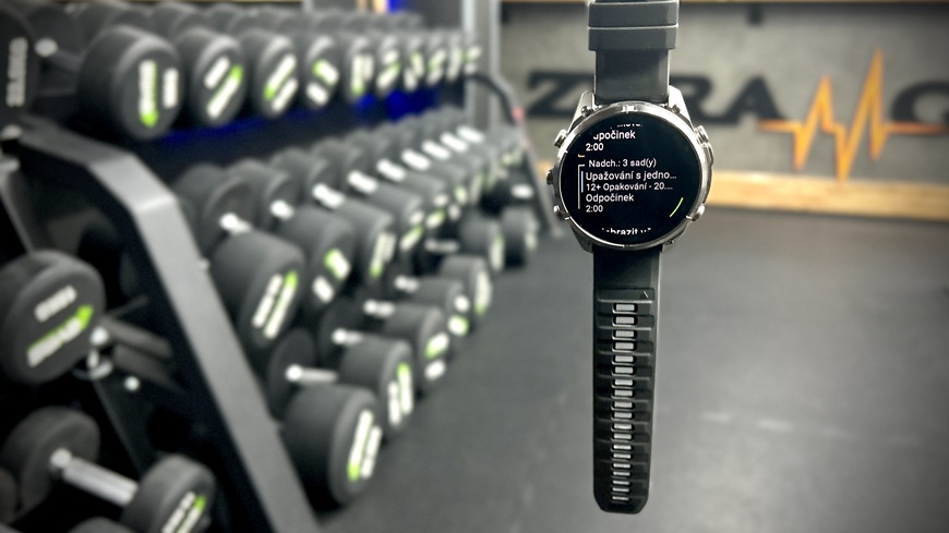Inteligentné hodinky Garmin fēnix® 9 Pro visiace v popredí stojana s činkami v posilňovni