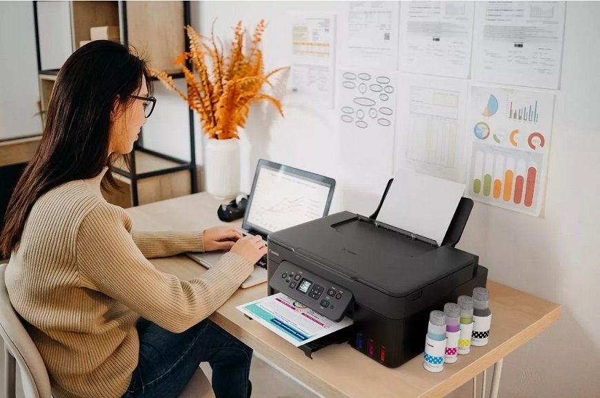 Ako vybrať tlačiareň? Žena pracuje na notebooku. Vedľa seba na stole má tlačiareň Canon PIXMA G3480.