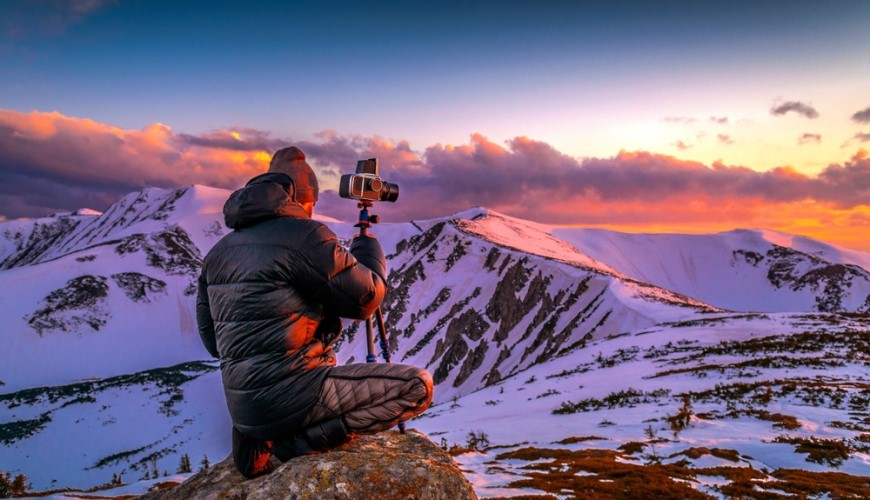Jak vybrat stativ na mobil, fotoaparát či kameru? Muž v horách fotografuje zasneženú krajinu s fotoapáratom na statíve.