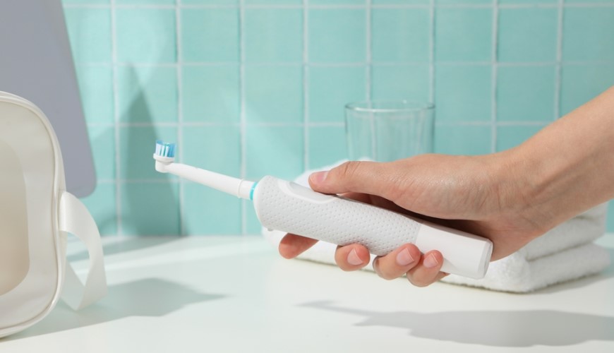 Ako vybrať elektrickú zubnú kefku? Ženská ruka drží elektrickú zubnú kefku s okrúhlou hlavicou, detailný pohľad v kúpeľni v pozadí je uterák a pohár s vodou