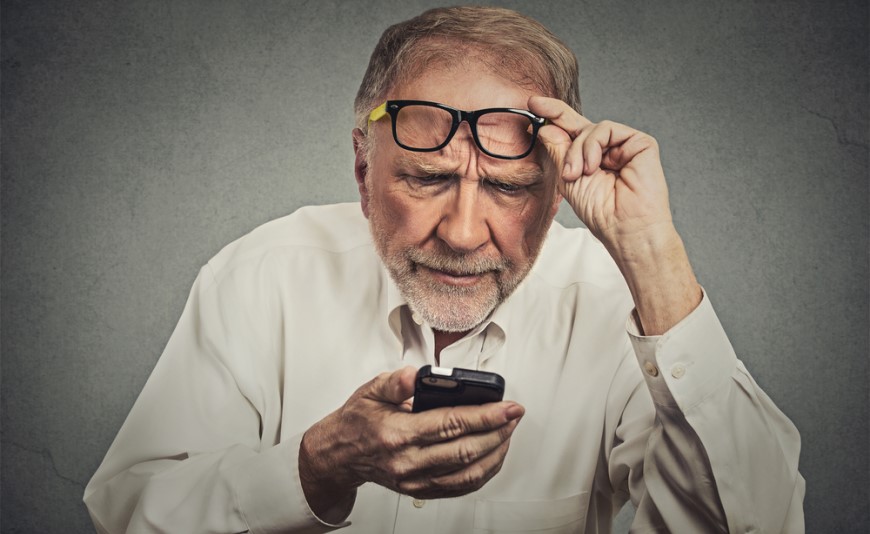 Najlepšie mobily pre seniorov Starší muž hľadí na tlačidlový telefón. Druhou rukou si drží zdvihnuté okuliare.