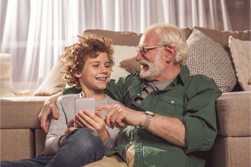 Najlepšie mobily pre seniorov Starší muž sedí na zemi vedľa vnuka. Opierajú sa o pohovku a smejú sa niečomu na vnukovom mobile.