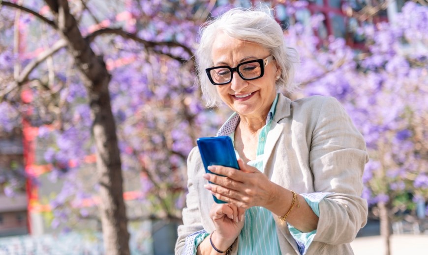 Najlepšie mobily pre seniorov Staršia žena stojí v parku a číta si správu na telefóne. V pozadí sú rozkvitnuté stromy.