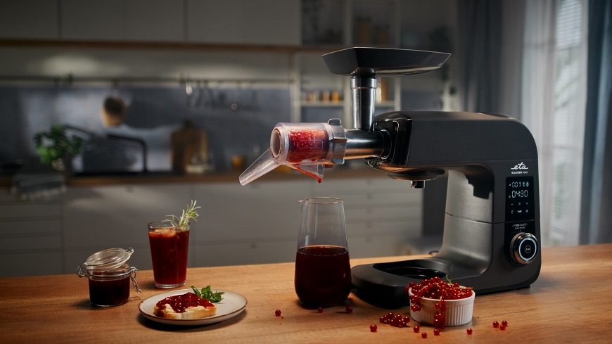 Čierny kuchynský robot ETA Gratus Kuliner II Max s nástavcom na lisovanie ovocia a zeleniny v akcii pri príprave ríbezľovej šťavy, s výsledným produktom v džbáne, pohári a zaváraninovom pohári s džemom.