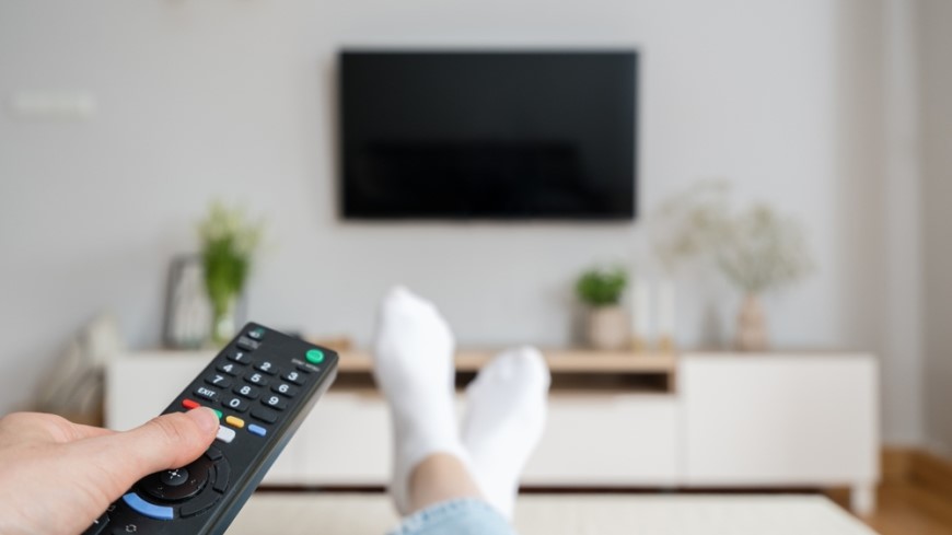 Ako pripojiť TV k Wi-Fi? ovládač od TV v ruke namierený na TV na stene, vyložené nohy na gauči