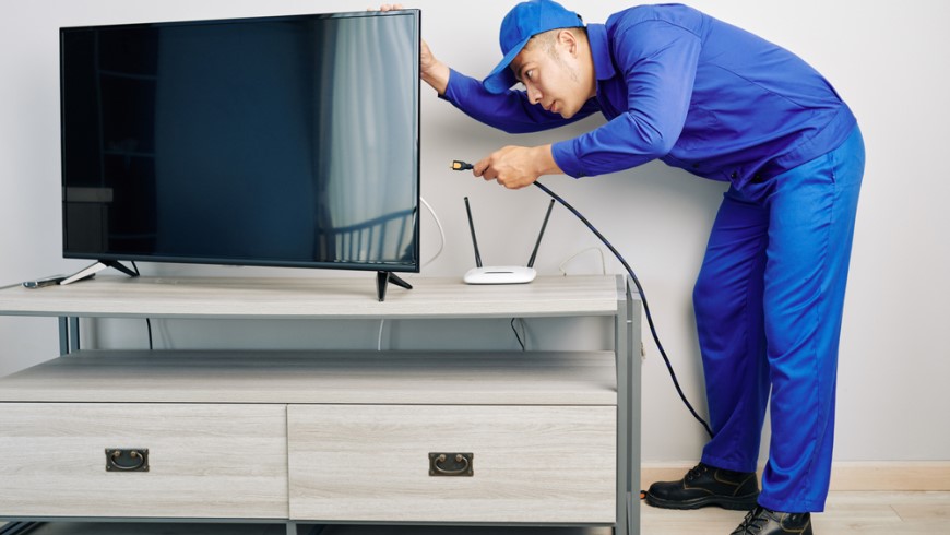 Ako bojovať so zlým signálom televízora? muž v modrých montérkach a šiltovke sa zohýba a zapája anténu do zadnej časti televízora, ktorý je položený na stolíku pod televízor