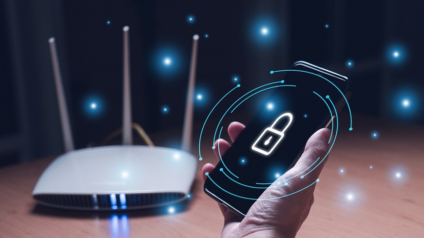 Detail ruky, ktorá drží smartfón so symbolom zámku na displeji, v pozadí je router na stole. Na obrázku sú aj svetlé modré body, akoby žiariace diódy.
