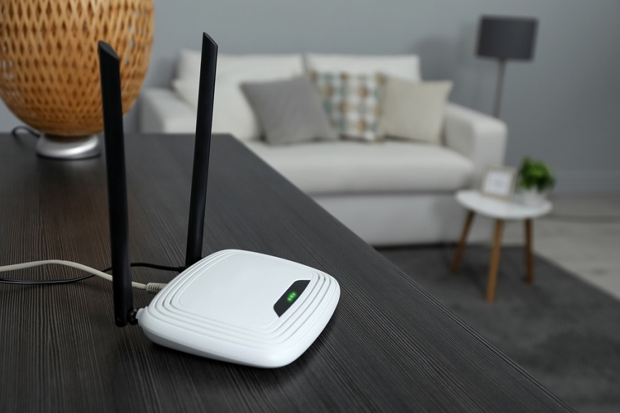 biely router s čiernymi anténami je položený na tmavom stole v obývačke s bielou pohovkou, na stole za routerom je oranžová dizajnová lampa
