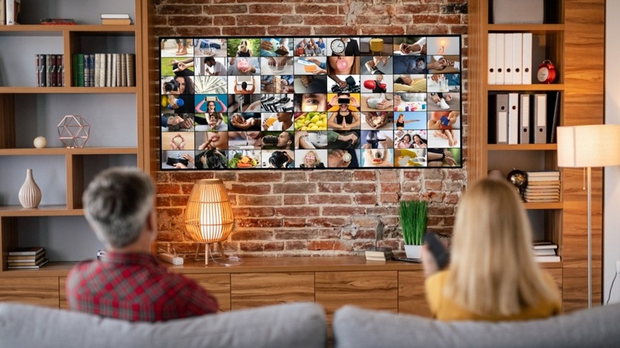Ako nainštalovať TV na stenu? Muž a žena sedia na gauči v obývačke a sledujú TV zavesený na stene plný obrazov