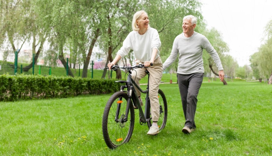 staršia žena sa vezie na elektrobicykli v parku a starší muž jej pridŕža sedadlo