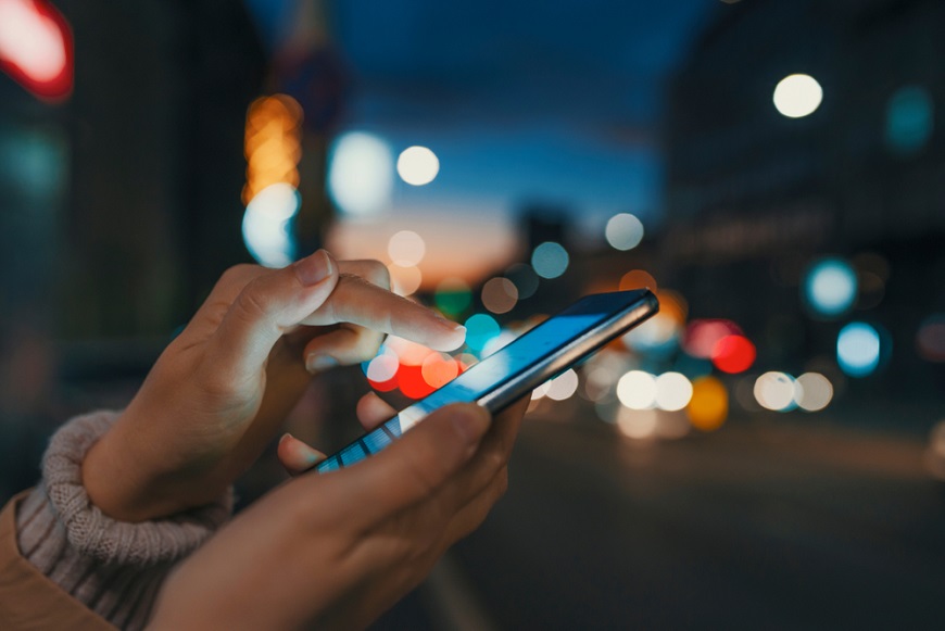Ako reštartovať mobil? detail rúk ovládajúcich smartfón, v pozadí rušná ulica po zotmení
