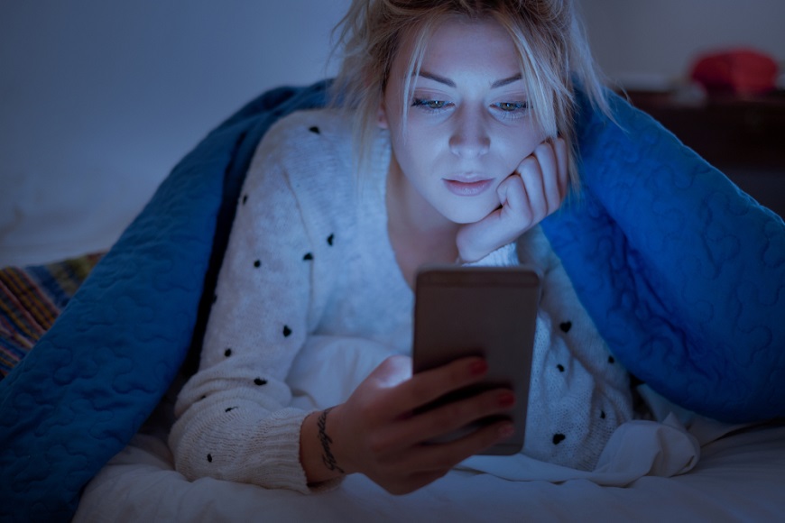 Na čo sú dobré okuliare proti modrému svetlu? žena pozerá v posteli a prítmí do mobilu