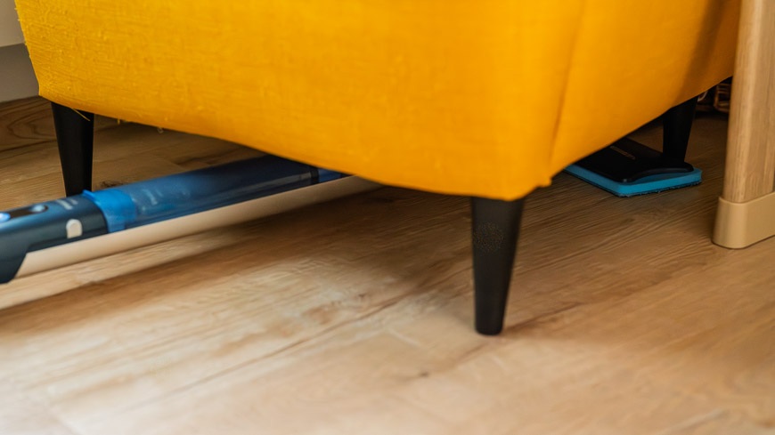  Mop Philips OneUp zasunutý pod žltým kreslom – položený na podlahe.