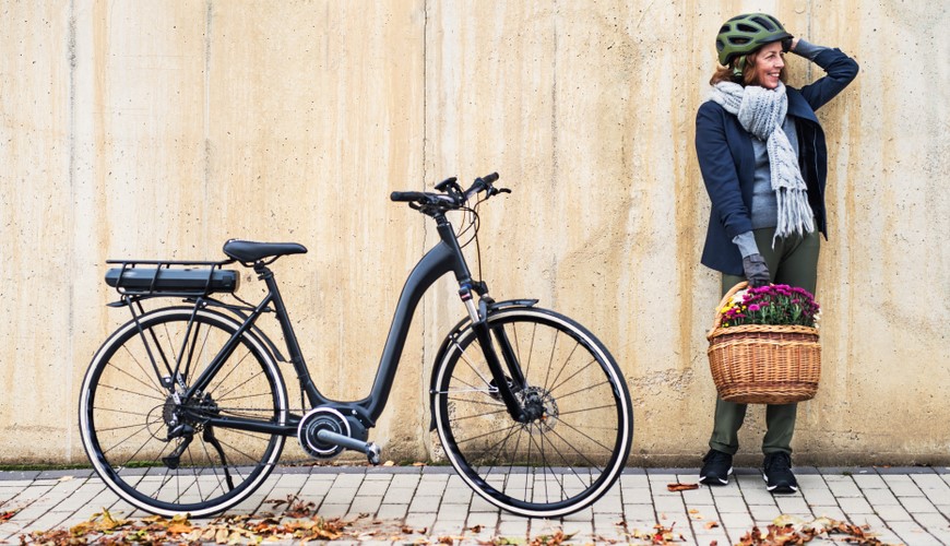 Ako vybrať elektrobicykel? žena s prilbou a košíkom plným kvetov stojí vedľa bicykla