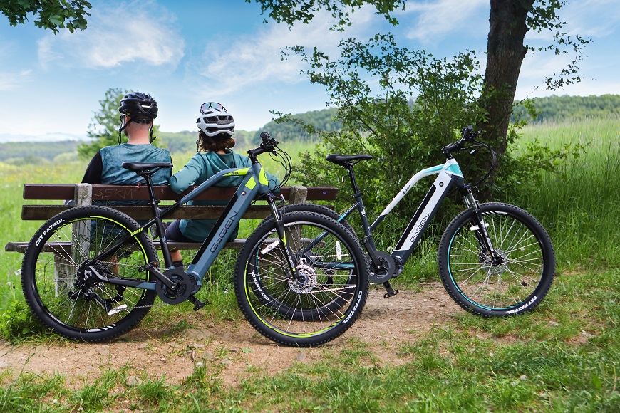 Ako vybrať elektrobicykel? muž a žena v cyklistickej výstroji sedia na lavičke v prírode, vedľa nich je bicykel, pohľad spoza nich