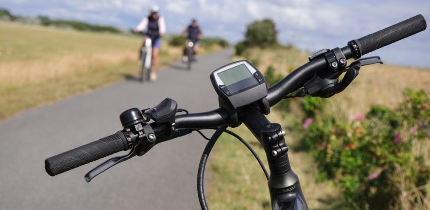 Ako vybrať elektrobicykel? detailný záber na tachometer na riadidlách bicykla v popredí, rozostrení cyklisti na cyklotrase v pozadí