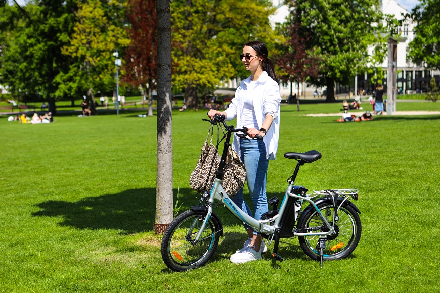 Ako vybrať elektrobicykel? žena v okuliaroch prechádza cez park po tráve so skladacím elektrobicyklom po boku