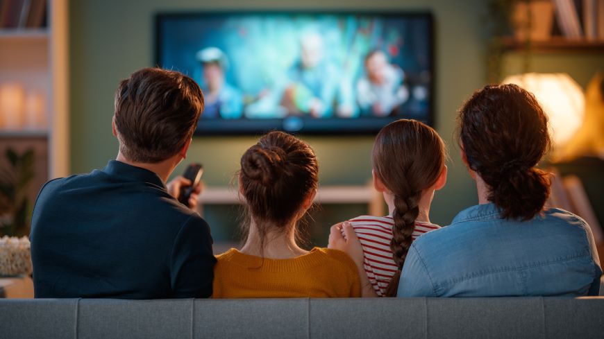 Ako ovládať televízor mobilom? Muž a žena s dvomi deťmi sedia pred televízorom – vidíme ich zozadu. Muž ovláda televízor diaľkovým ovládačom.