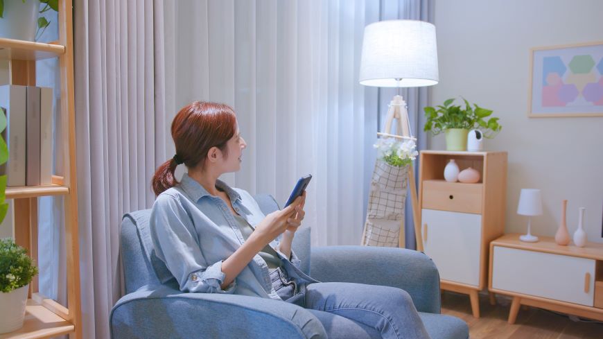 Ako ovládať televízor mobilom? Žena sedí v modrom kresle medzi nábytkom a hľadí vpravo na lampu. Tá svieti. Žena drží v ruke mobilný telefón.