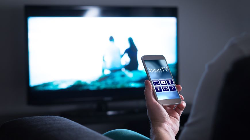 Ako ovládať televízor mobilom? Mobilný telefón s aplikáciou Smart TV na displeji. V pozadí zapnutý televízor.