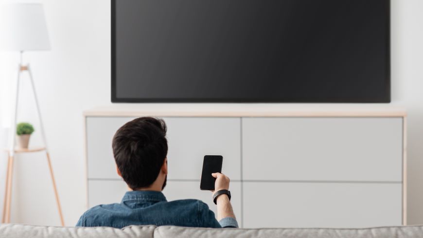 Ako ovládať televízor mobilom? Muž sedí na pohovke pred vypnutým televízorom a mieri naň mobilným telefónom.