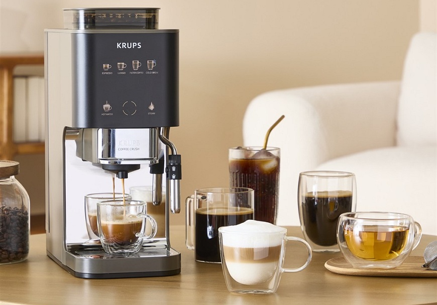 Kávovar Krups Coffee Crush Experience na stole pripravuje kávu. Okolo ďalšej šálky a pohára s kávovými špecialitami a čajom.