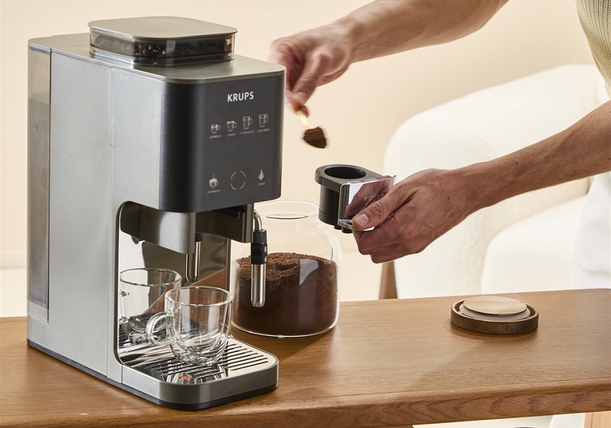  Kávovar Krups Coffee Crush na stole. Žena naberá do zásuvky na kávu lyžičkou mletú kávu zo sklenenej dózy.