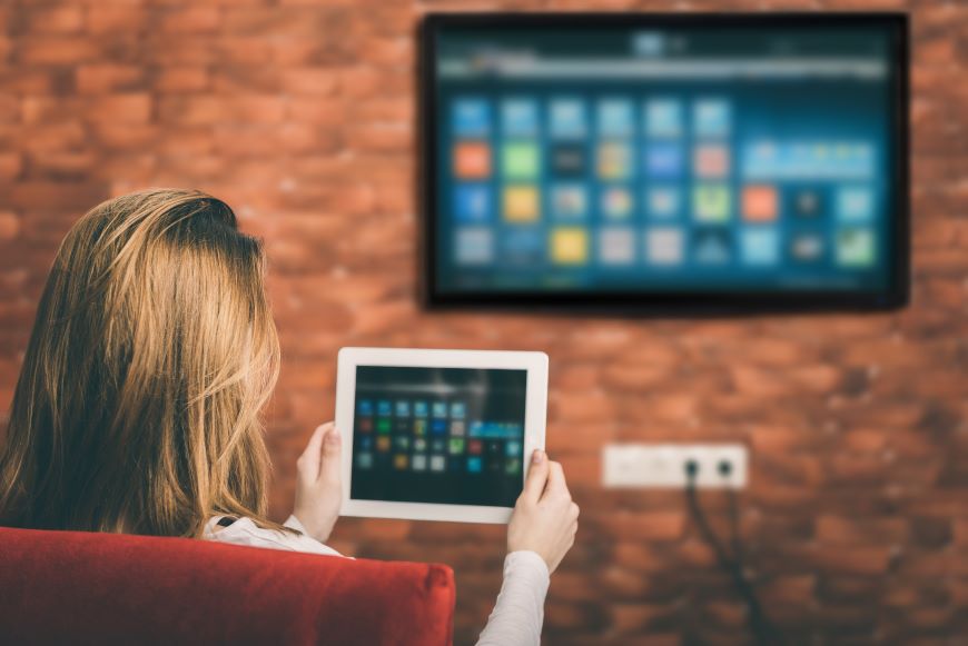 Ako prepojiť tablet s televízorom? Žena sedí na pohovke pred televízorom a drží pred sebou tablet. Na TV sa zrkadlí to isté, čo na displeji