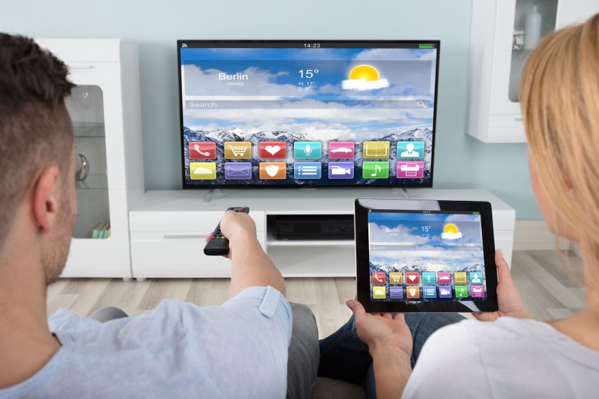 Ako prepojiť tablet s televízorom? Muž so ženou sedia na pohovke pred televízorom. Muž má k TV natiahnutú ruku s diaľkovým ovládačom. Žena pred sebou drží tablet, ktorého displej sa zrkadlí na TV.