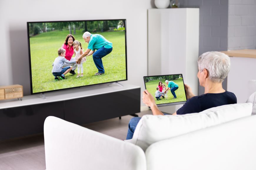 Ako prepojiť tablet s televízorom? Staršia žena sedí na pohovke pred TV a pozerá sa na tablet, ktorý drží v rukách. Na tablete je rovnaká fotografia rodiny s deťmi ako na TV.