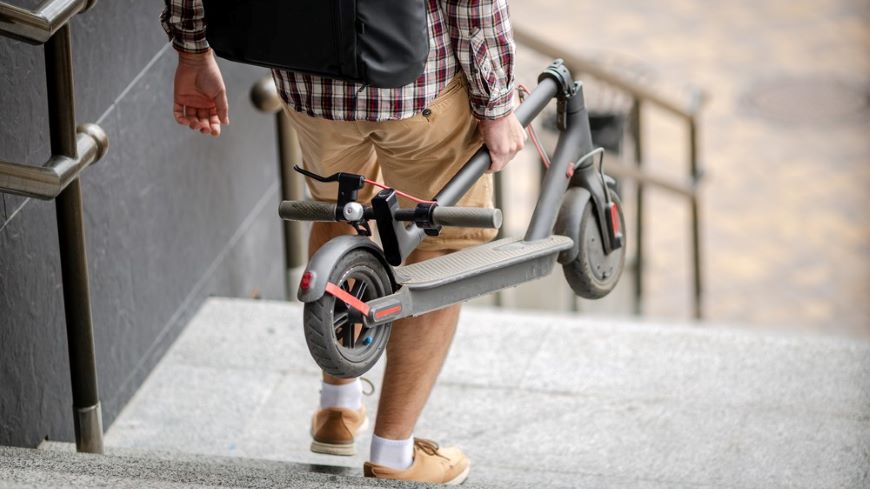 Ako vybrať elektrokolobežku? muž v krátkych nohaviciach a flanelovej košeli schádza po schodoch a v rukách nesie zloženú elektrokolobežku, pohľad zozadu