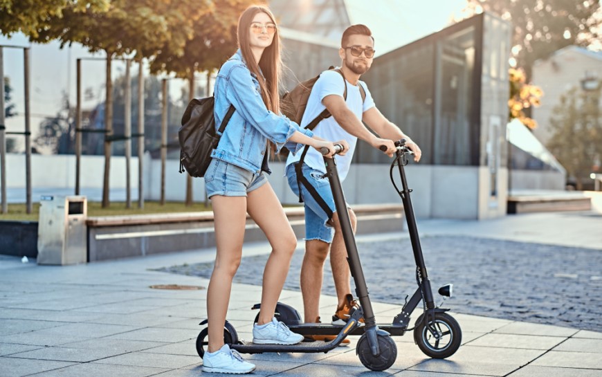 Ako vybrať elektrokolobežku? mladý muž a žena v letnom oblečení na kolobežkách v meste na námestí