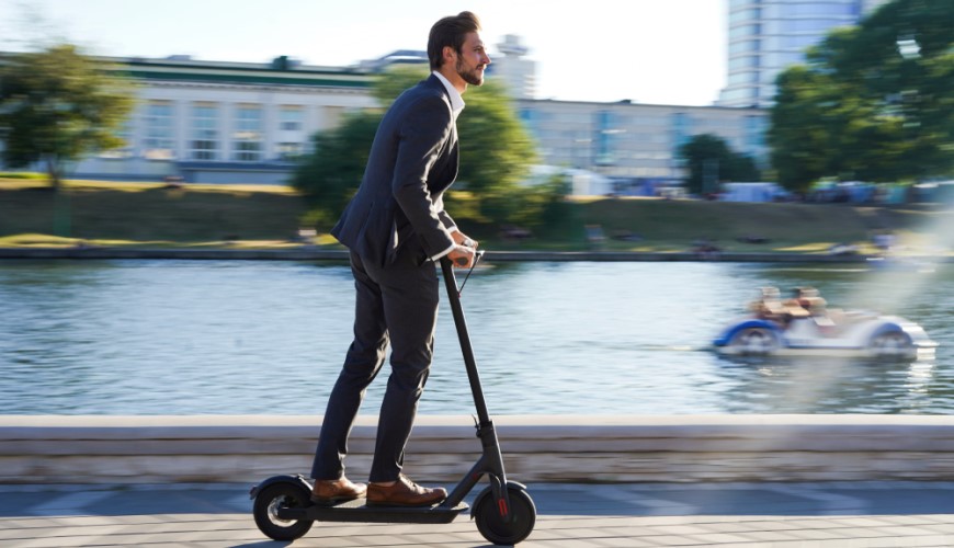 Ako vybrať elektrokolobežku? muž v obleku ide na kolobežke okolo fontány v meste