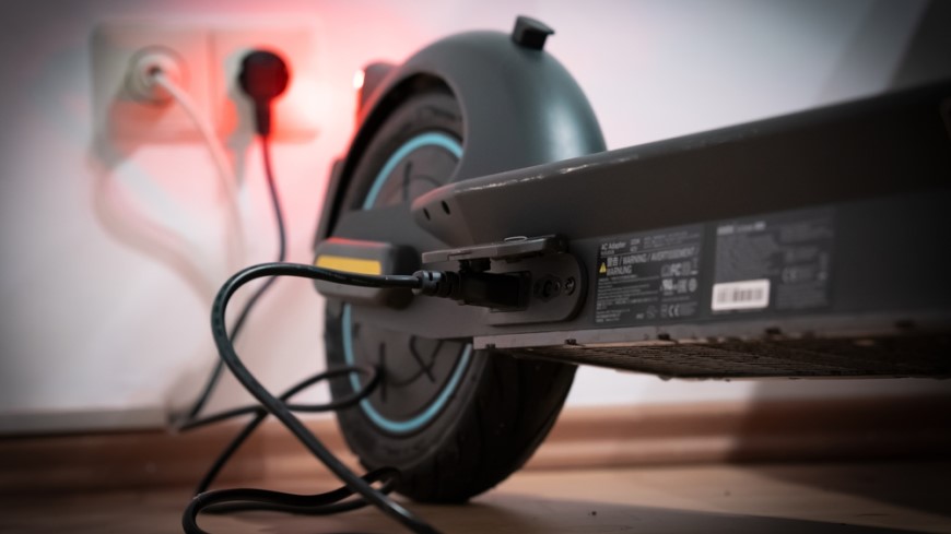 Ako vybrať elektrokolobežku? detailný pohľad na nabíjanie elektrokolobežky cez kábel, v miestnosti je prítmie a priestor je osvetľovaný červenou diódou