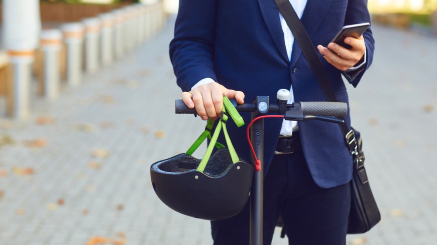 Ako vybrať elektrokolobežku? muž v obleku drží telefón v jednej ruke a v druhej riadidlá kolobežky, prilbu má zavesenú na riadidlách