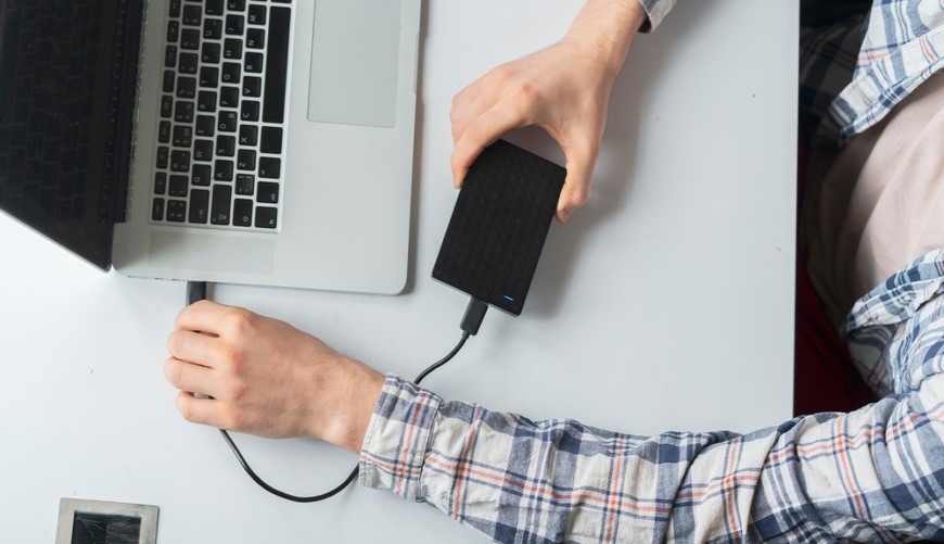 muž sediaci za pracovným stolom zapája externý disk do notebooku, pohľad zhora