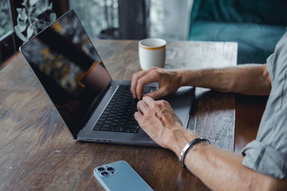 muž sedí v kaviarni za stolom a píše na macbooku, detailný záber na ruky, stôl a notebook