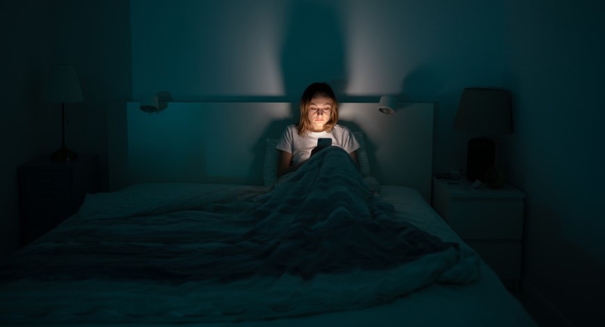 Ako vypnúť modré svetlo na iPhone je tma, žena sedí prikrytá perinou v posteli v spálni a pozerá do mobilu, z ktorého vyžaruje silné svetlo