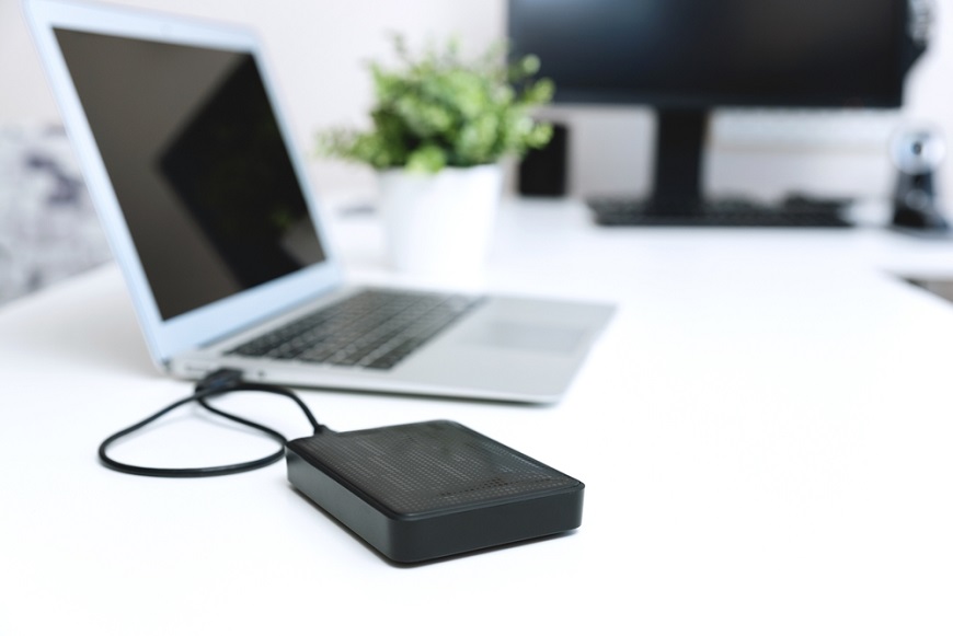 Ako preniesť dáta do nového telefónu otvorený laptop s pripojeným externým diskom na bielom PC stole, v rozostrenom pozadí je monitor a izbový kvet