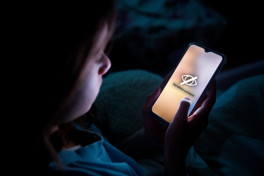 Ako nastaviť rodičovskú kontrolu na inteligentných zariadeniach? Dievča leží v noci posteli a do tváre jej svieti displej mobilu, na ktorom je symbol pre sensitive content.