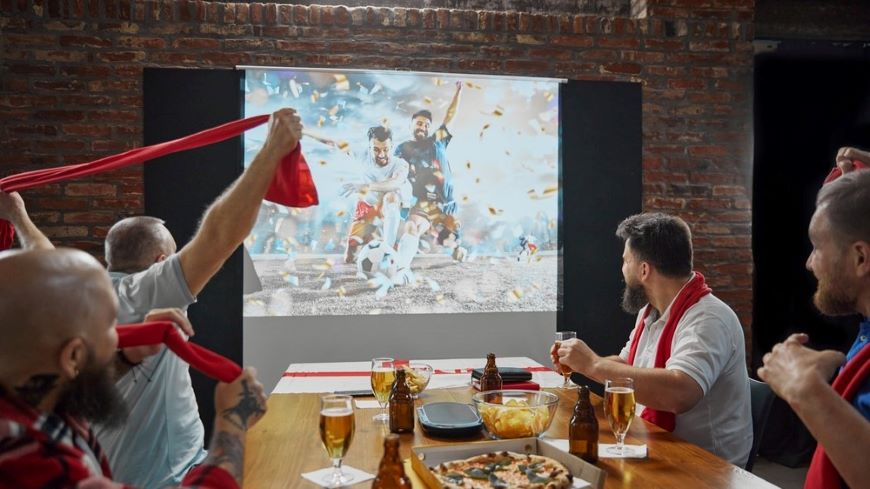 Ako vybrať projektor? skupina mužov s červenými šálmi fandí futbalu, ktorý je premietaný na plátne v pohostinstve, sedia za stolom s občerstvením