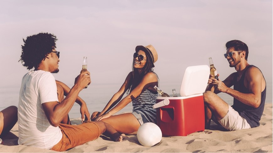 Ako vybrať chladničku na cesty dvaja muži so ženou sedia na pláži a popíjajú drink vychladený v absorpčnej chladničke