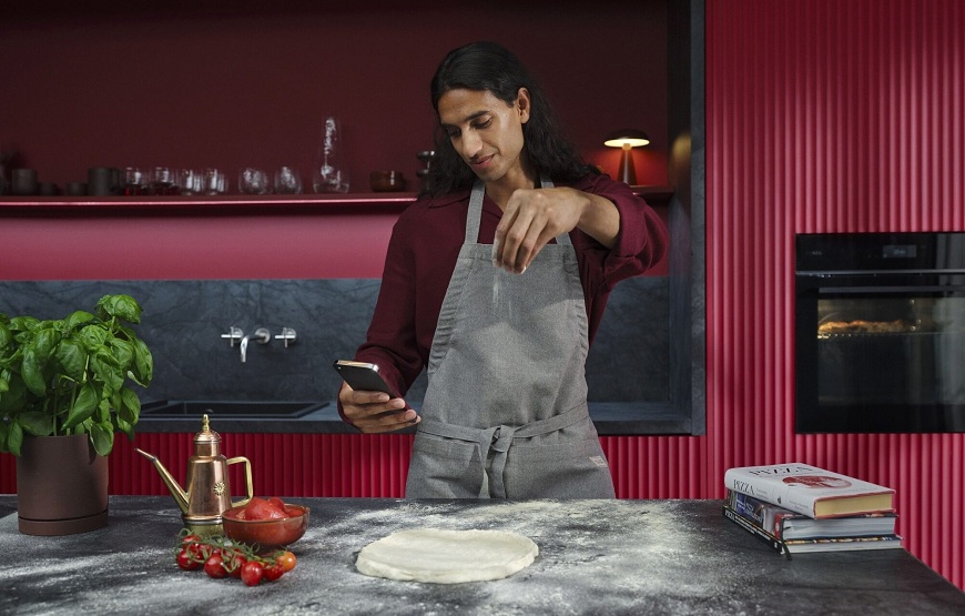 Nová rúra AEG PizzaExpert: Pizza ako z Neapola už za 2,5 minúty Muž pripravuje pizzu a pozerá sa do mobilného telefónu. V pozadí je červená kuchyňa s rúrou AEG.