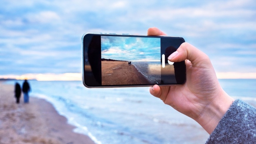 Smartphone fotografujúci pár v pozadí, idúci po pláži vedľa možského príboja.