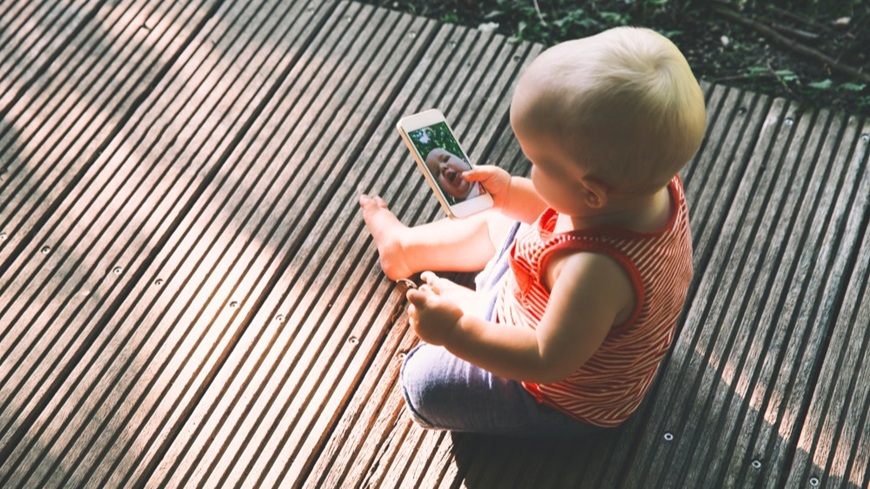 Batoľa sediaca na verande a držiaca v ruke mobilný telefón so spustenou fotoaplikáciou