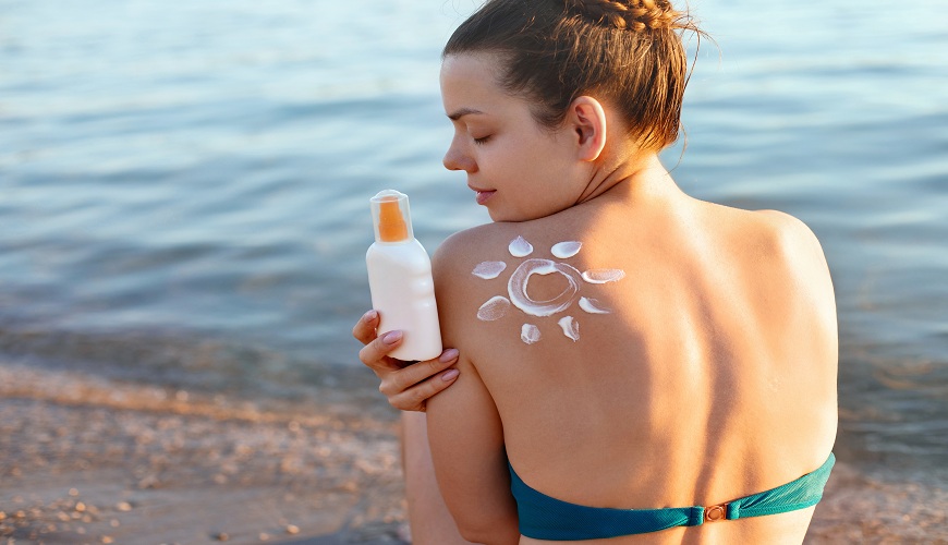 Soľ, pot a SPF: Ako v lete chrániť pleť aj vlasy bez zbytočného zaťaženia Mladá žena sedí na pláži a natiera sa opaľovacím krémom