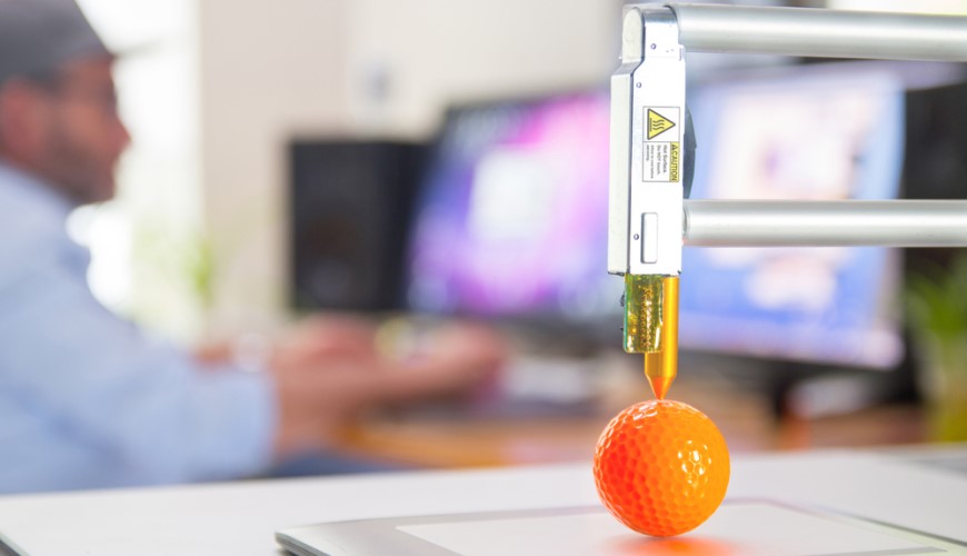 Ako vybrať 3D tlačiareň? 3D tlačiareň vytvára oranžovú golfovú loptičku, v pozadí je muž pracujúci na PC