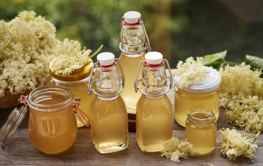 Ako pripraviť domáci sirup? Chuť leta ukrytá v každej kvapke Bazový sirup v flašiach a kvetmi položený na drevennom stole.
