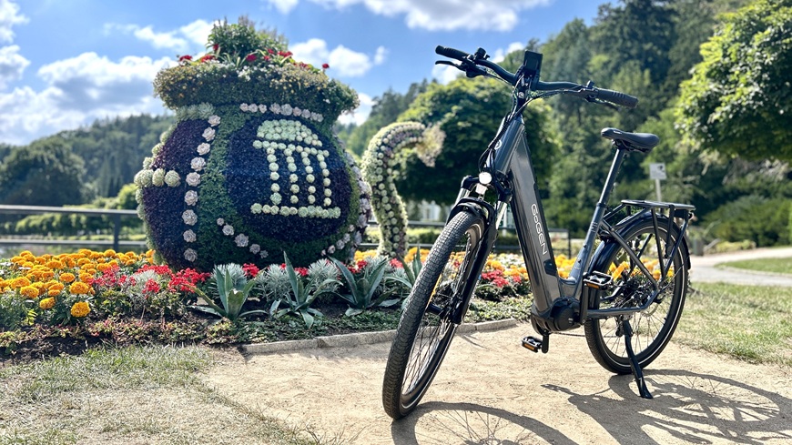 Recenzia: Elektrobicykel GoGEN Aspen MX 720 sa postará o to, aby vaše cyklistické túžby nič nebrzdilo Elektrobicykel GoGEN Aspen MX 720 v šedo-čiernom prevedení opretý o stojan pred slnkom zaliatym kvetinovým záhonom